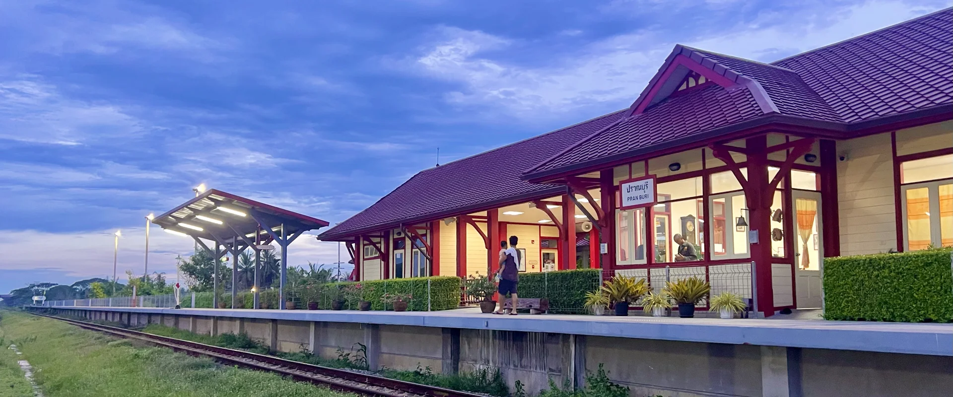 Pran Buri Railway Station’s red-and-cream building, illuminated platform and bilingual station sign at dusk.