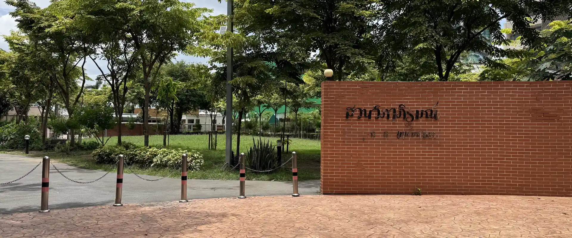 Vibhaphirom Park entrance sign on a red brick wall beside trees and lawn in Chom Phon, Bangkok.