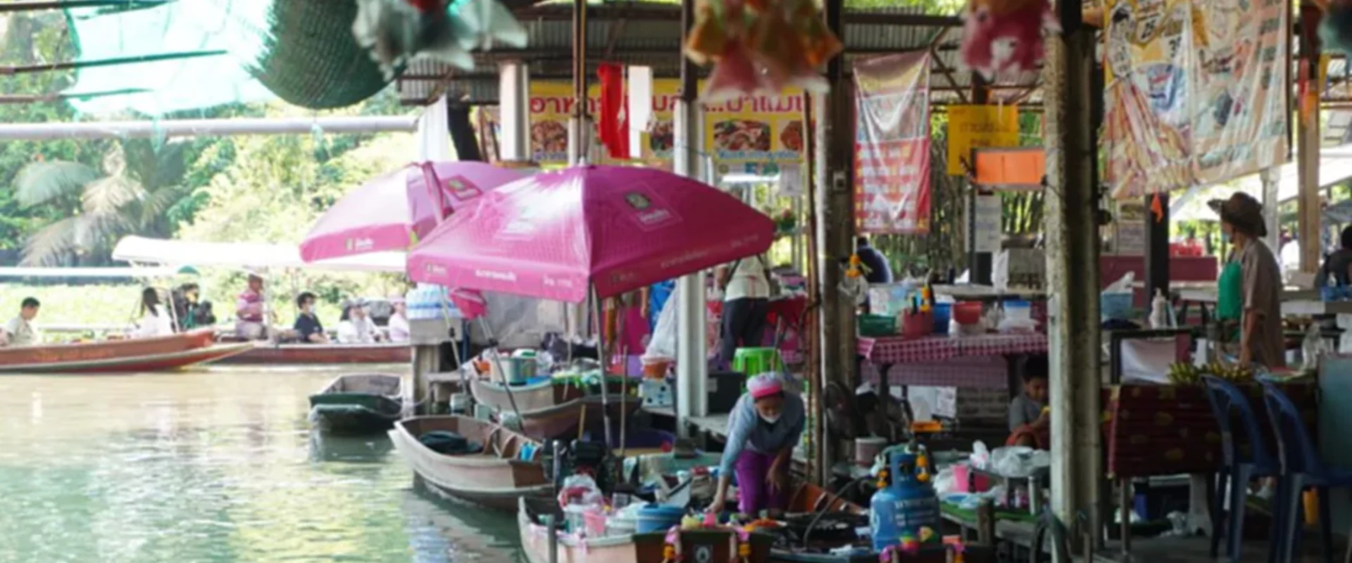 Boat vendors and pink umbrellas beside the covered waterside stalls at Wat Saphan Floating Market, Bangkok.